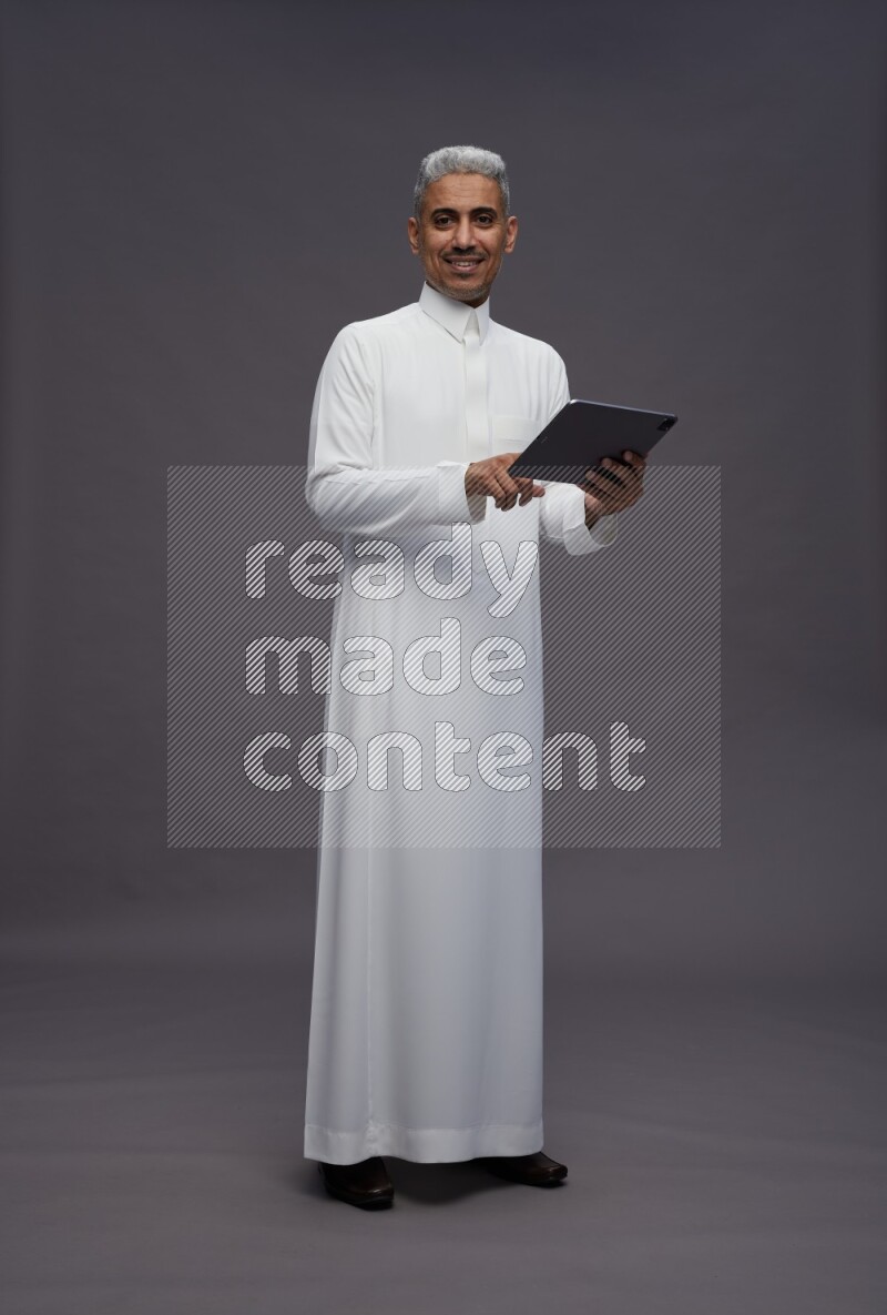 Saudi man wearing thob standing working on tablet on gray background
