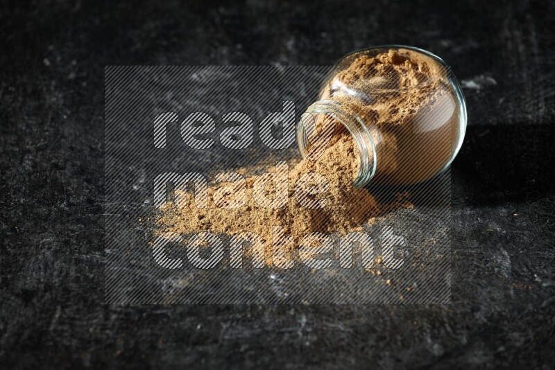 A flipped glass spice jar full of allspice powder and powder spilled out of it on a textured black flooring