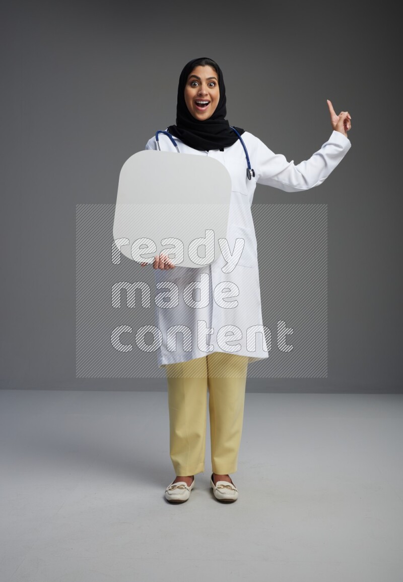 Saudi woman wearing lab coat with stethoscope standing holding social media sign on Gray background