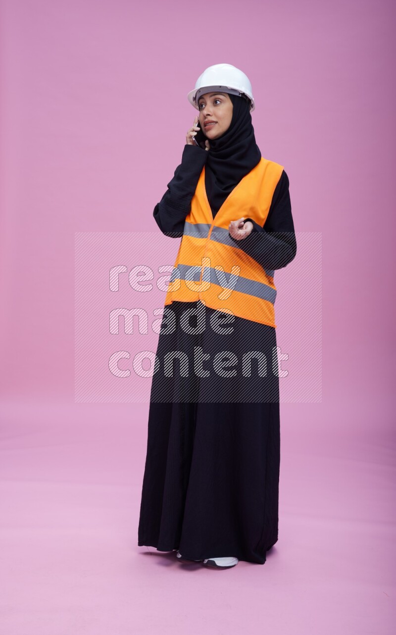 Saudi woman wearing Abaya with engineer vest and helmet standing talking on phone on pink background