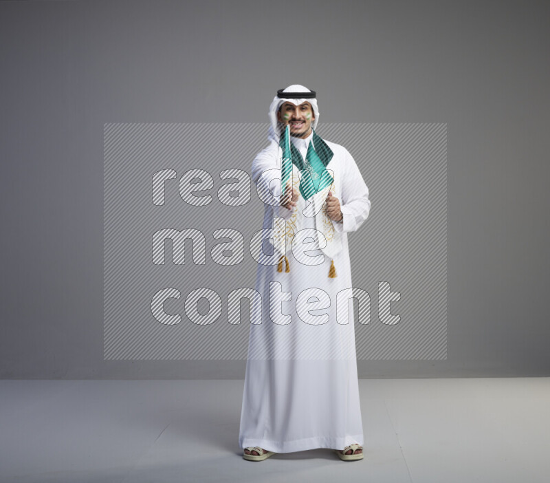 A saudi man standing wearing thob and white shomag with face painting and saudi flag scarf and holding small saudi flag on gray background