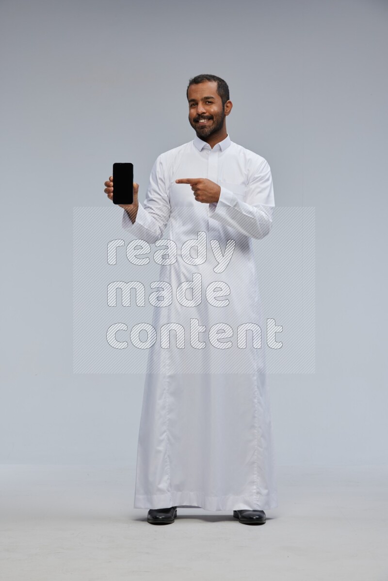 Saudi man wearing Thob standing showing phone to camera on Gray background
