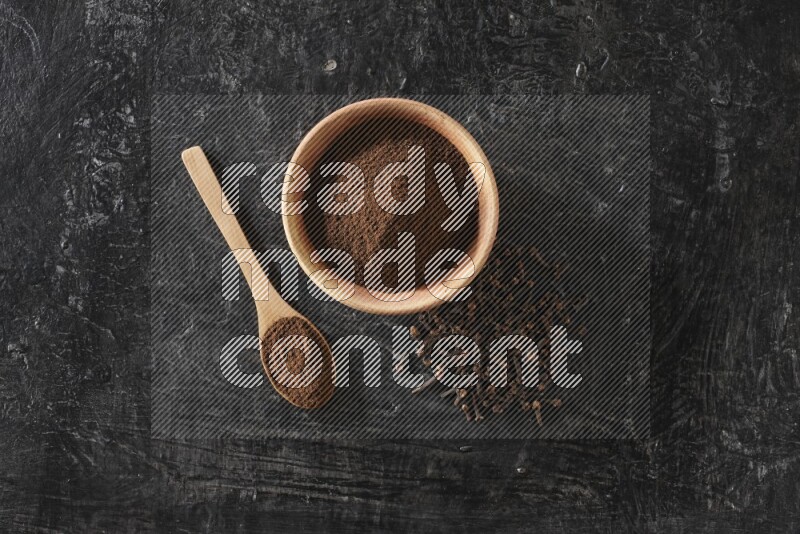 A wooden bowl and a wooden spoon full of cloves powder with spreaded cloves on a textured black flooring