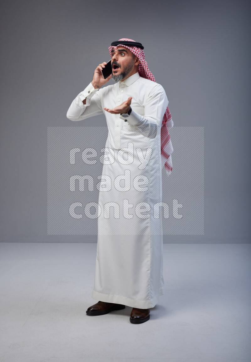 Saudi man Wearing Thob and red Shomag standing talking on phone on Gray background
