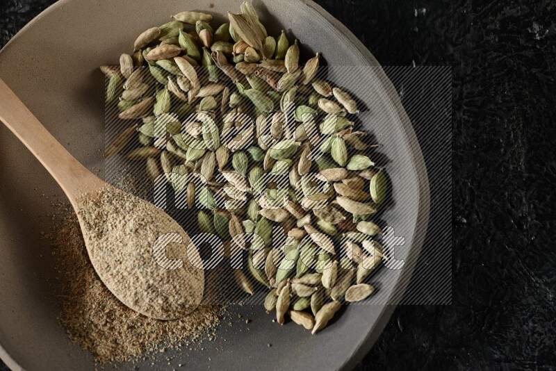 A plate filled with cardamom seeds and a wooden spoon full of cardamom powder on a textured black flooring