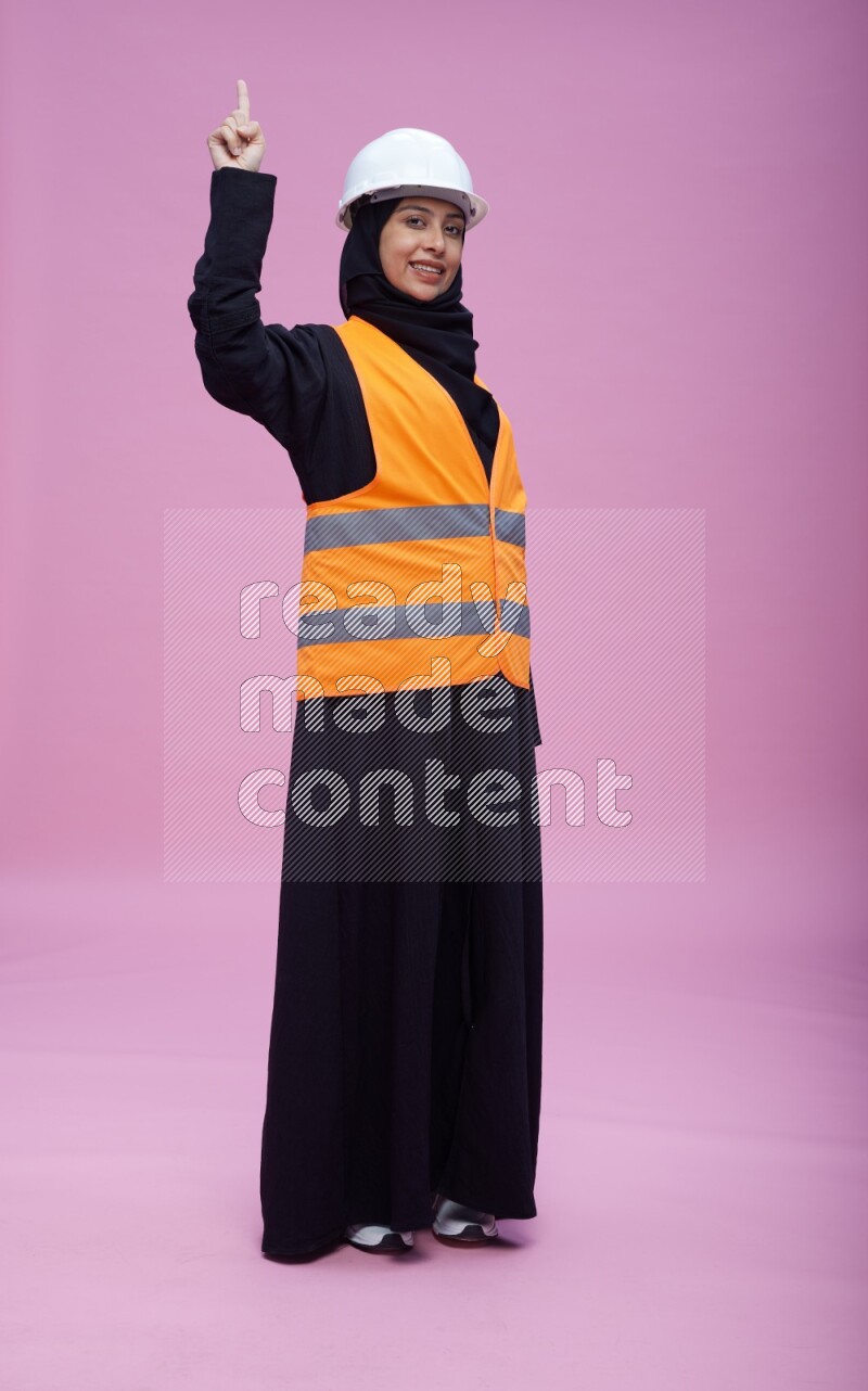 Saudi woman wearing Abaya with engineer vest and helmet standing interacting with the camera on pink background