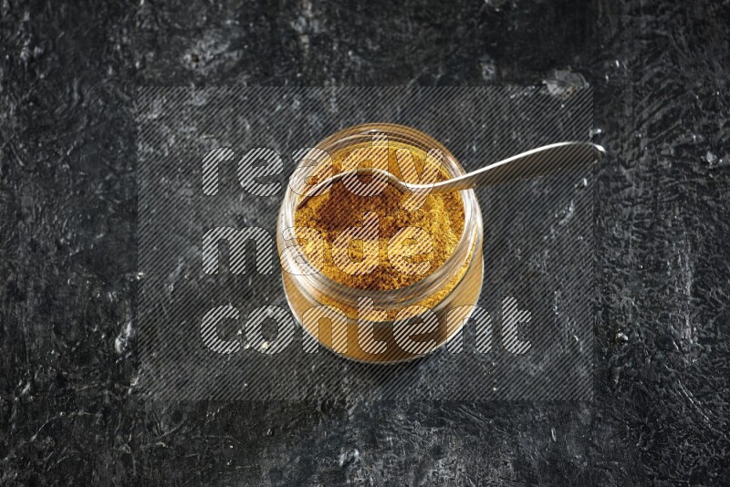 A glass jar and a metal spoon full of turmeric powder on a textured black flooring