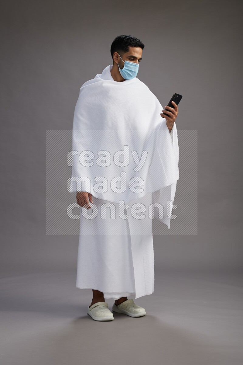 A man wearing Ehram with face mask Standing texting on phone on gray background