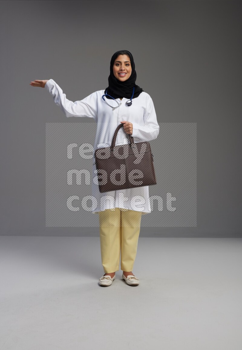 Saudi woman wearing lab coat with stethoscope standing holding bag on Gray background