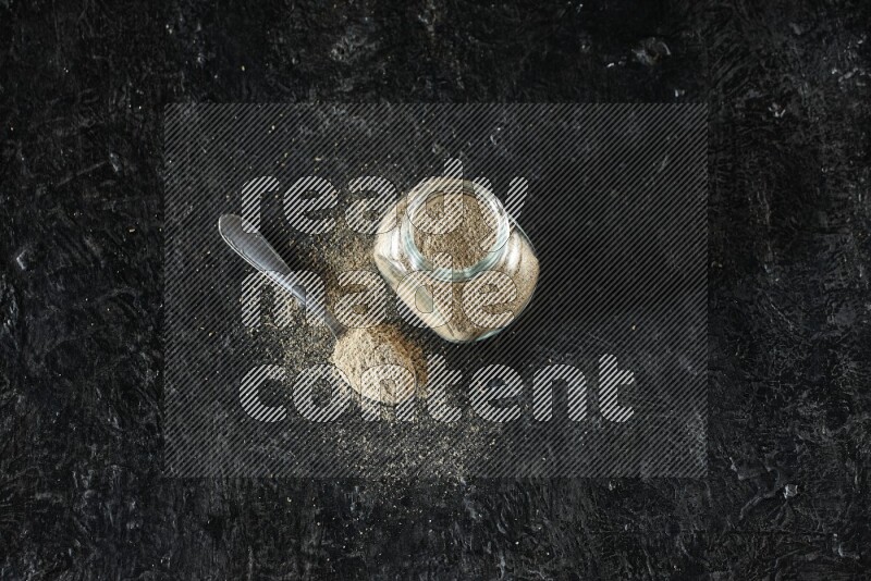 وعاء زجاجي للتوابل وملعقة معدنية ممتلئان ببودرة الهيل على خلفية سوداء