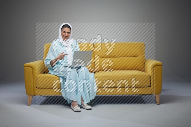 A Saudi woman sitting on a yellow Sofa With her Laptop Pointing wearing Blue Abaya