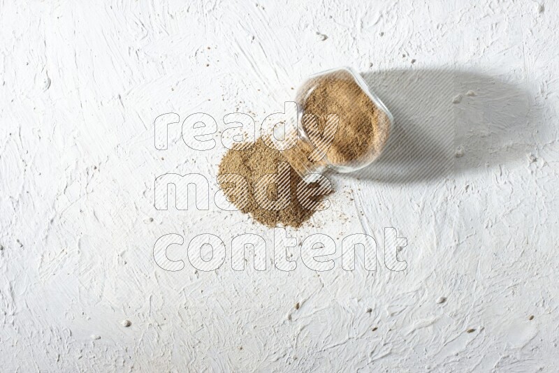 A flipped glass spice jar full of cumin powder and powder spilled out on textured white flooring