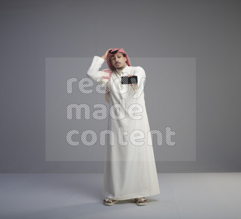 رجل سعودي يرتدي ثوب ابيض وشماغ احمر يشير الى هاتفه باتجاه الكاميرا