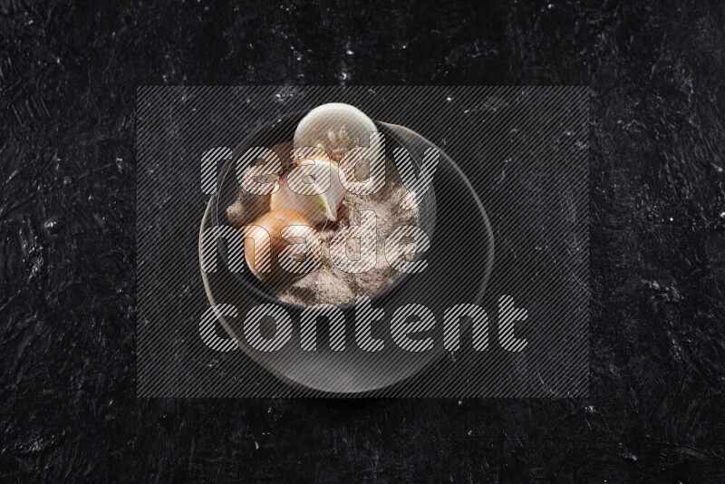 A black pottery bowl full of onion powder and some onion all on a pottery plate on black background