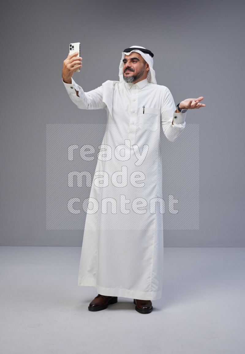 Saudi man Wearing Thob and white Shomag standing taking selfie on Gray background