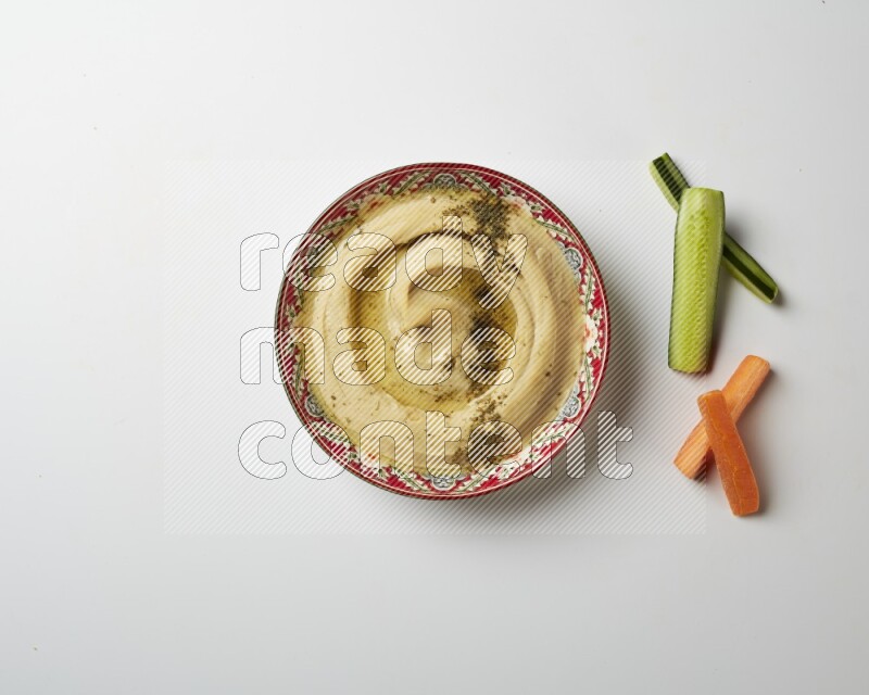 Hummus in a red plate with patterns garnished with zaatar & sumak on a white background