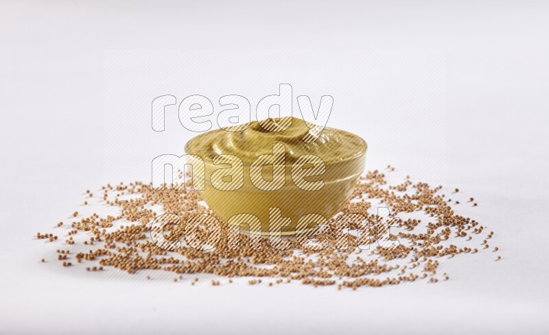 A glass bowl full of mustard paste with mustard seeds underneath on white flooring