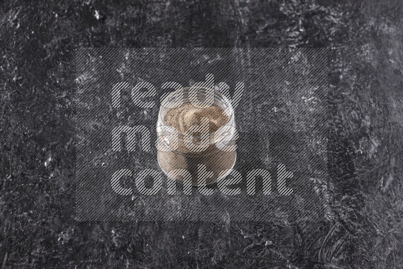 A glass jar full of black pepper powder on a textured black flooring