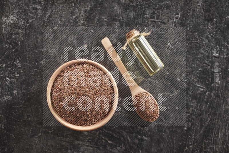 A wooden bowl and spoon full of flaxseeds and a glass bottle of flaxseeds oil on a textured black flooring