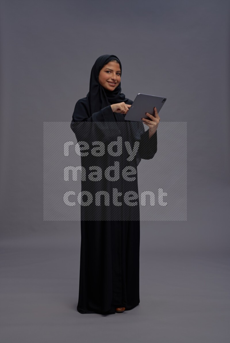 Saudi woman wearing Abaya standing working on tablet on gray background