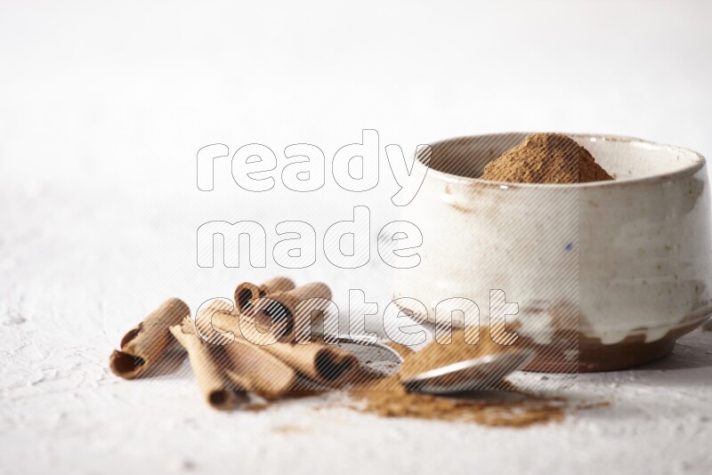 Ceramic beige bowl full of cinnamon powder and a metal spoon with cinnamon sticks next of it on a textured white background