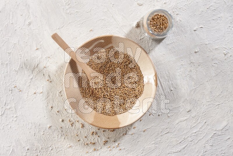 A beige pottery plate full of mustard seeds and a wooden spoon in it with a glass jar filled with the seeds on a textured white flooring