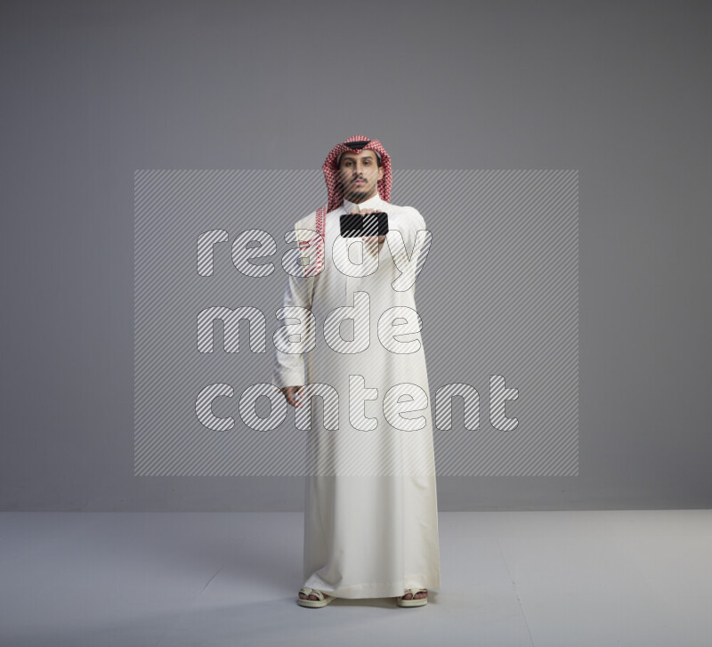 رجل سعودي يرتدي ثوب ابيض وشماغ احمر يشير الى هاتفه باتجاه الكاميرا