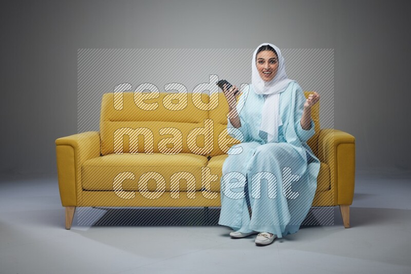 A Saudi woman wearing a light blue Abaya and white head scarf sitting on a yellow sofa and using her phone eye level on a grey background