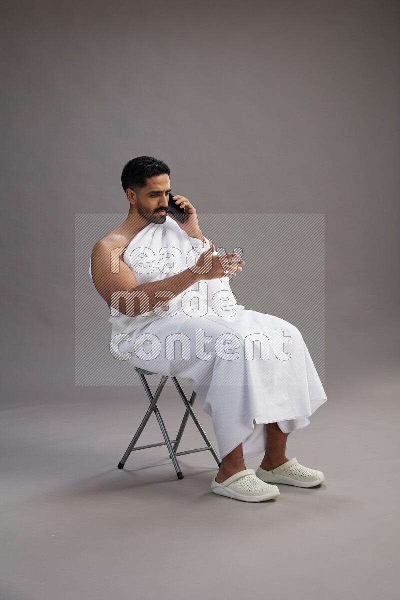 A man wearing Ehram sitting on chair talking on the phone on gray background
