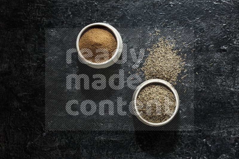 2 beige bowls, one full of cumin powder and the other full of seeds on a textured black flooring