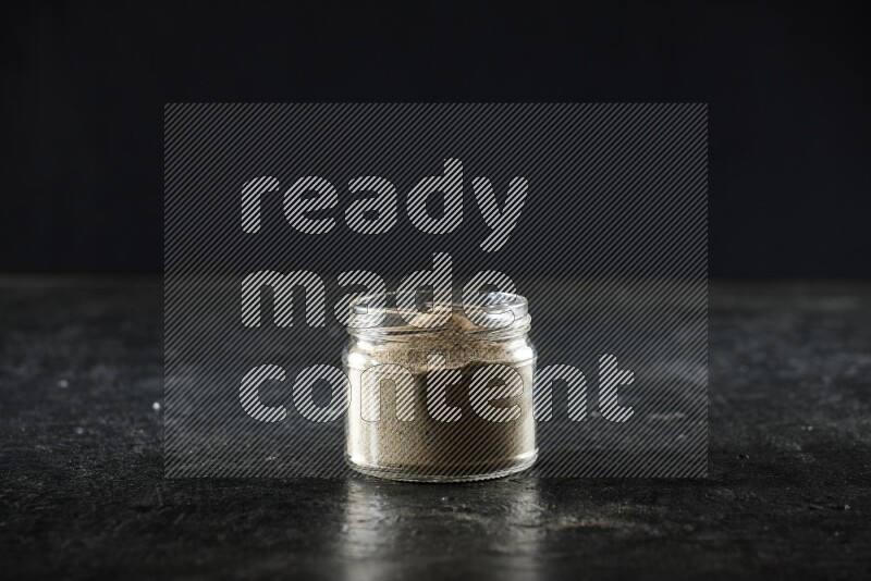 A glass jar full of cardamom powder on textured black flooring