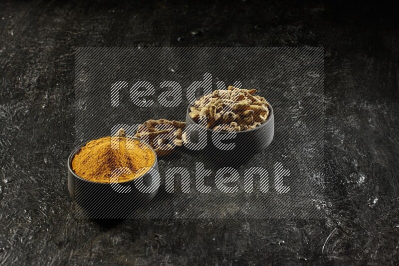 2 pottery black bowls, one full of turmeric powder and the other full of dried turmeric whole fingers on a textured black flooring