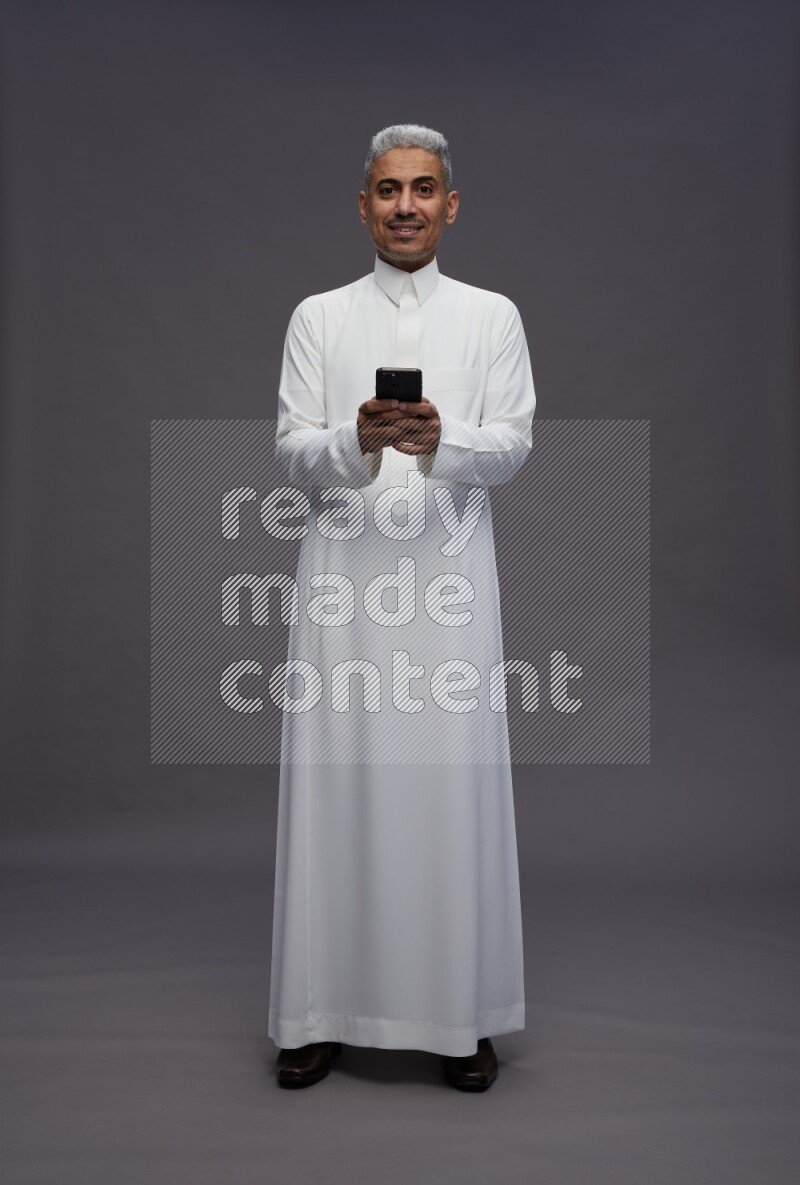 Saudi man wearing thob standing texting on phone on gray background