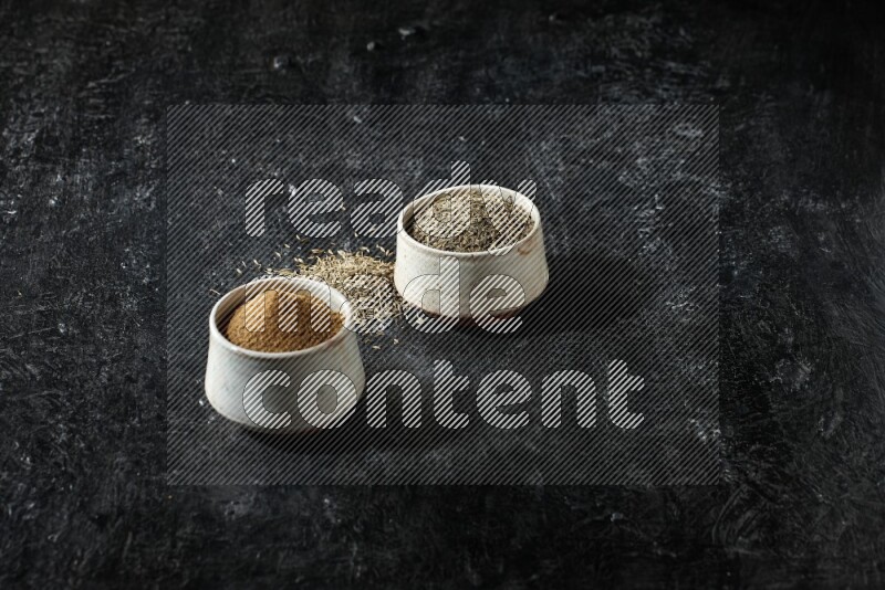 2 beige bowls, one full of cumin powder and the other full of seeds on a textured black flooring