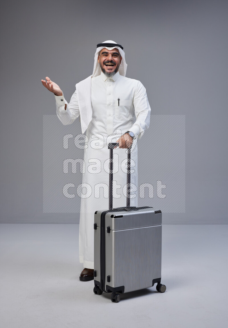 Saudi man wearing Thob and white Shomag standing holding Travel bag on Gray background