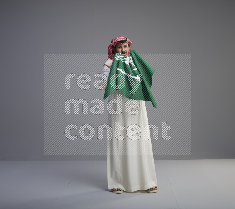 A saudi man standing wearing thob and red shomag holding big saudi flag on gray background