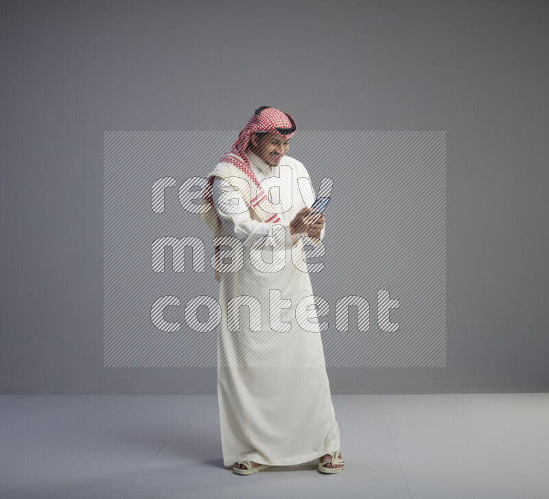 A  Saudi man standing wearing thob and red shomag texting on phone on gray background