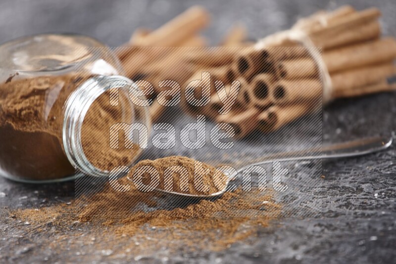 Herbal glass jar full cinnamon powder flipped and a metal spoon full of powder, cinnamon sticks stacked and bounded in the back on textured black background in different angles