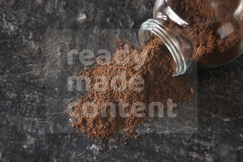 A flipped glass spice jar full of cloves powder and powder came out of it on textured black flooring