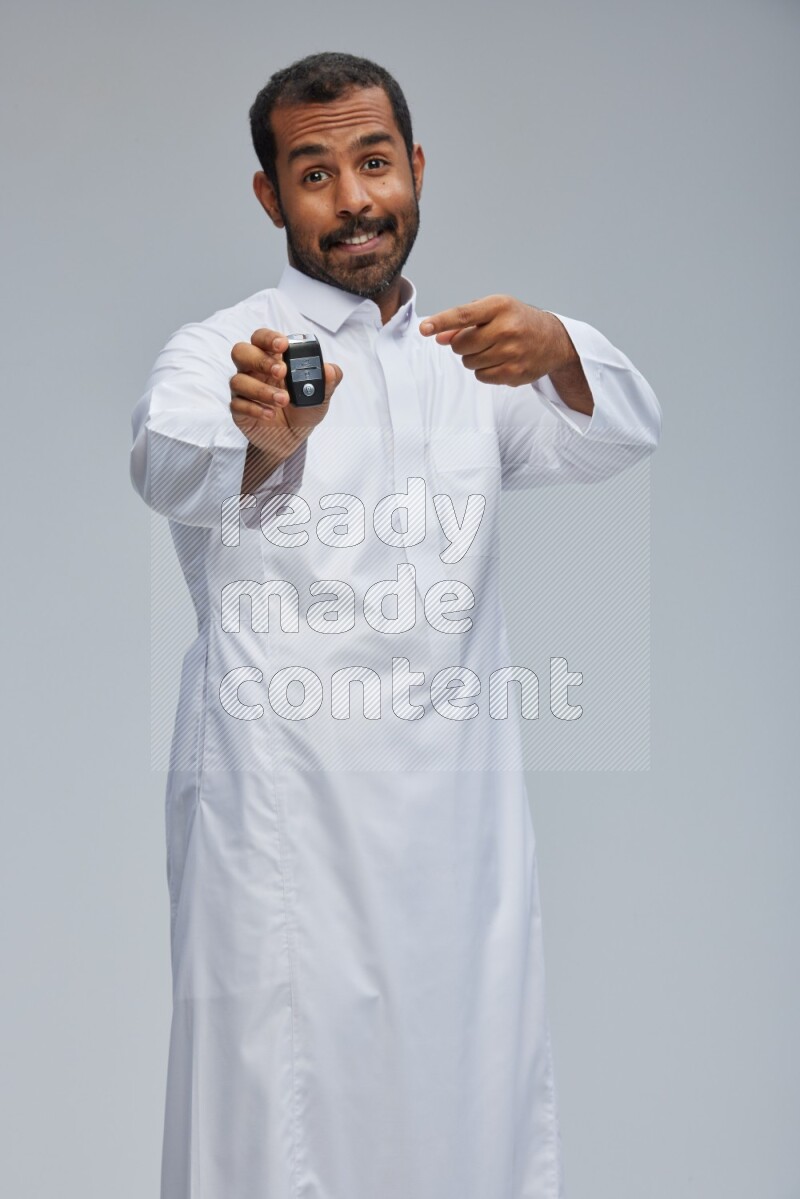 Saudi man wearing thob and shomag standing holding car key on gray background