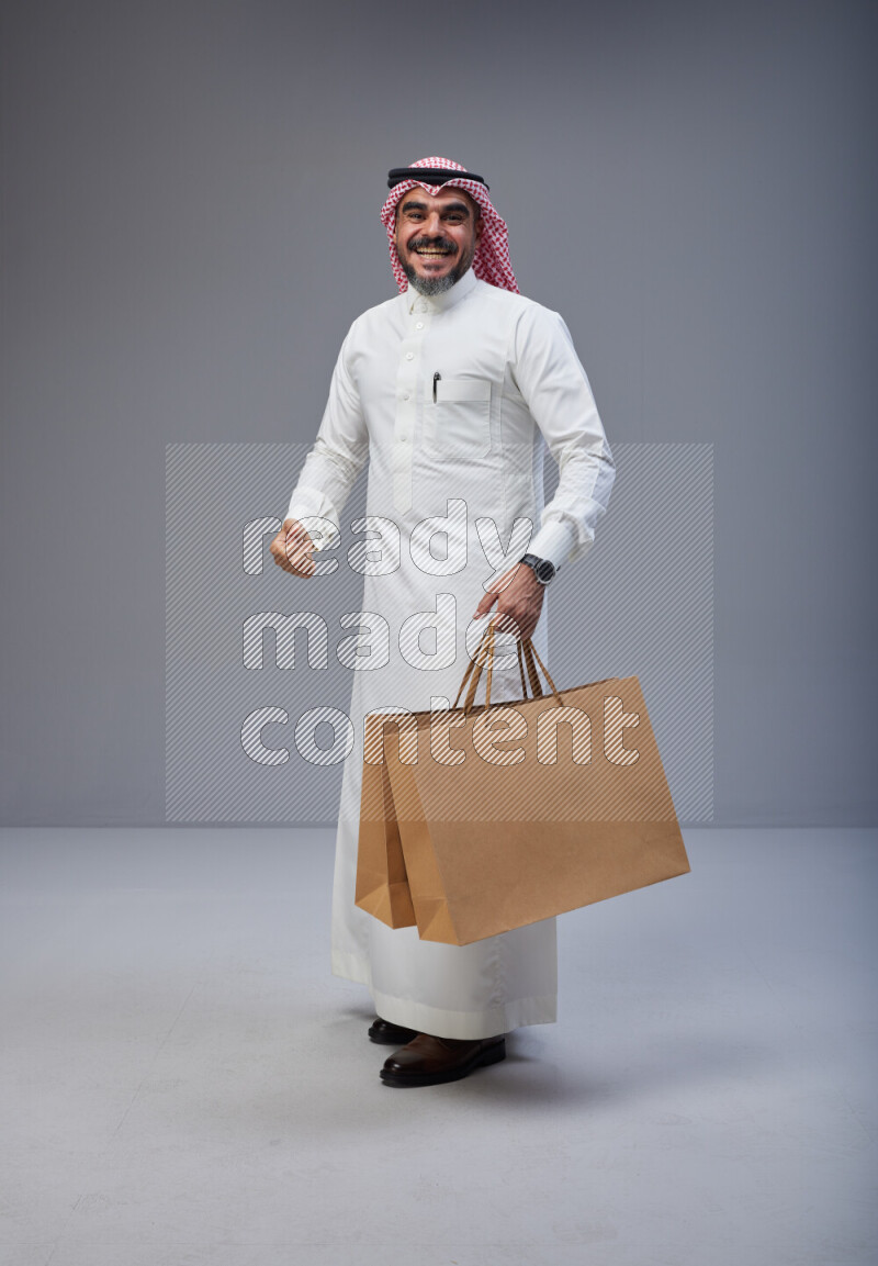 Saudi man Wearing Thob and red Shomag standing holding shopping bag on Gray background