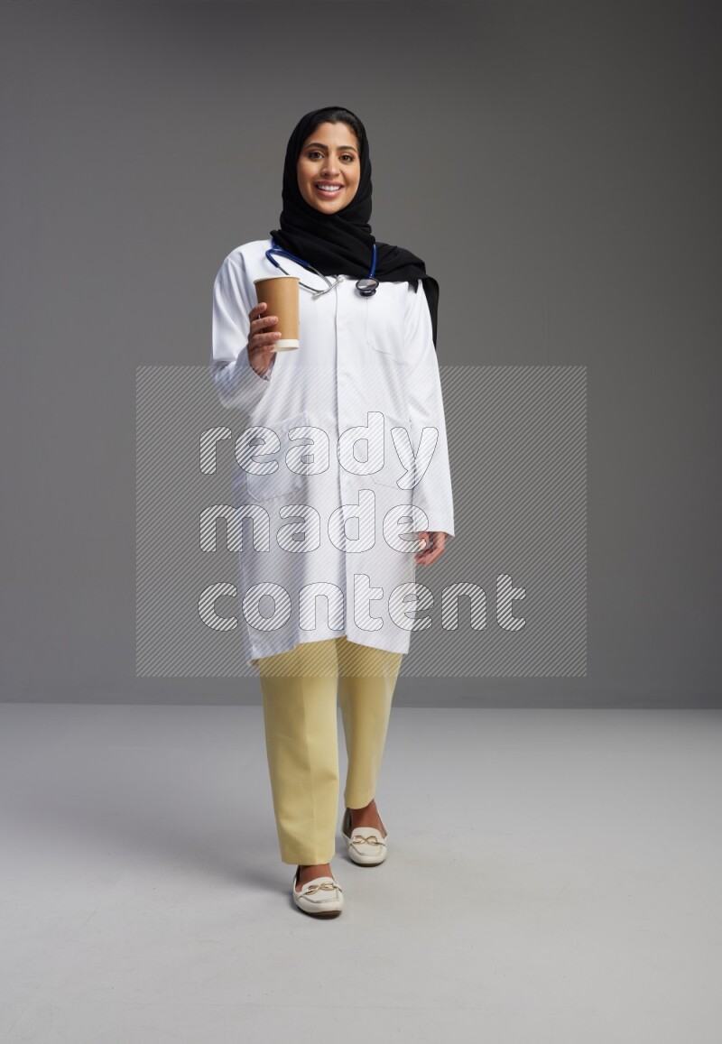 Saudi woman wearing lab coat with stethoscope standing holding paper cup on Gray background