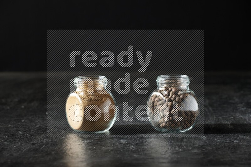 2 glass spice jars full of allspice powder and whole balls on a textured black flooring