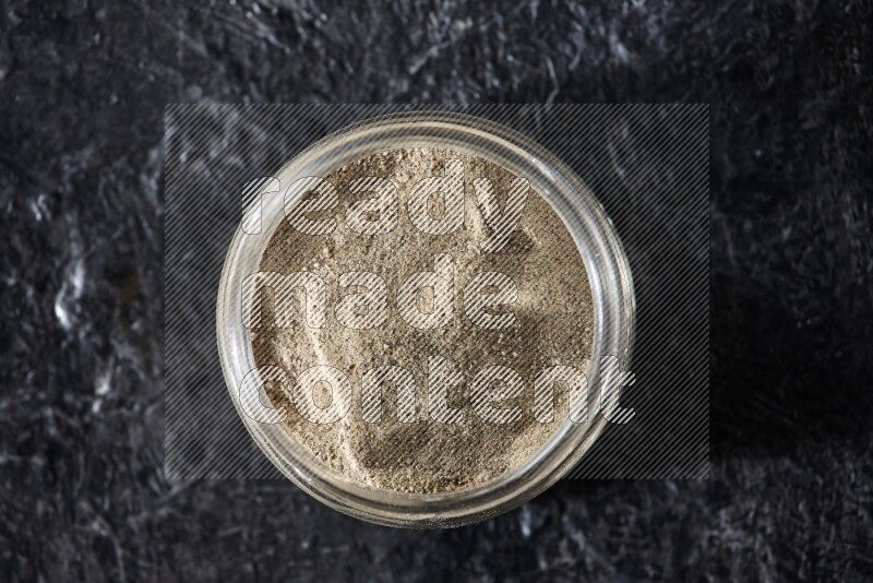 A glass jar full of white pepper powder on textured black flooring