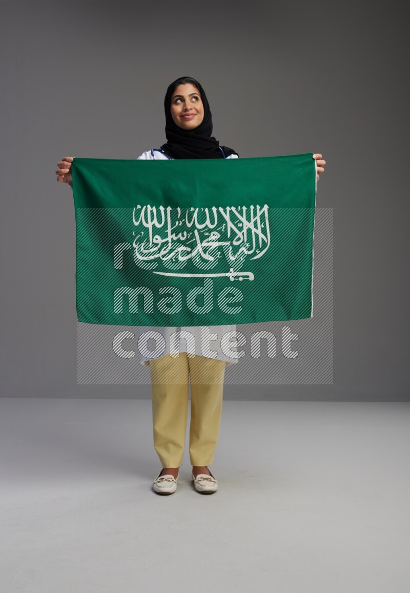 Saudi woman wearing lab coat with stethoscope standing holding Saudi flag on Gray background