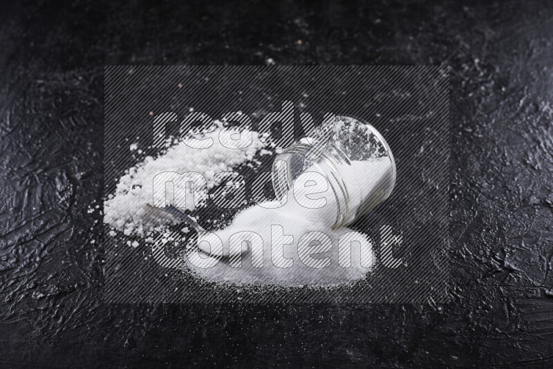 A glass jar full of table salt with some sea salt crystals beside it on a black background