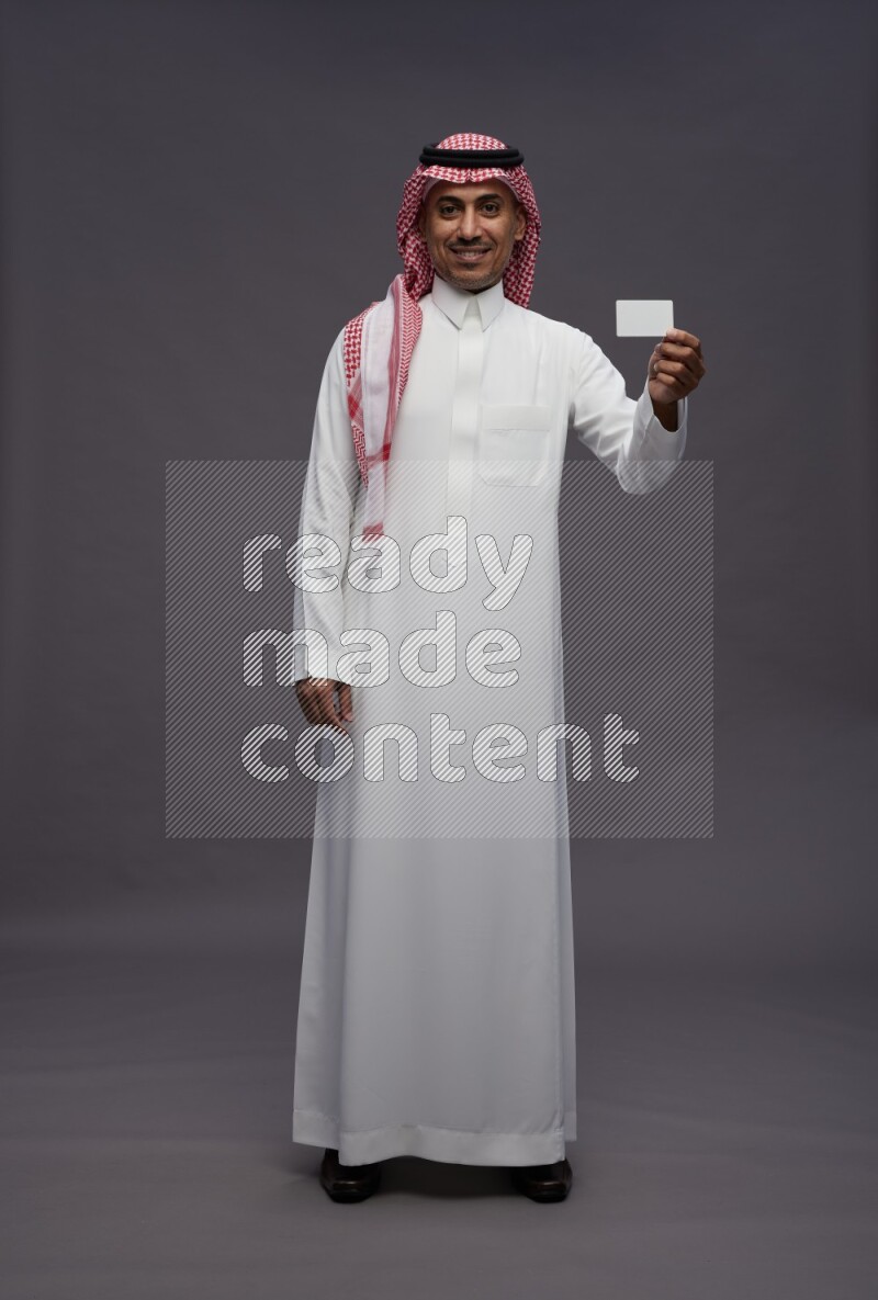 Saudi man wearing thob and shomag standing holding ATM card on gray background