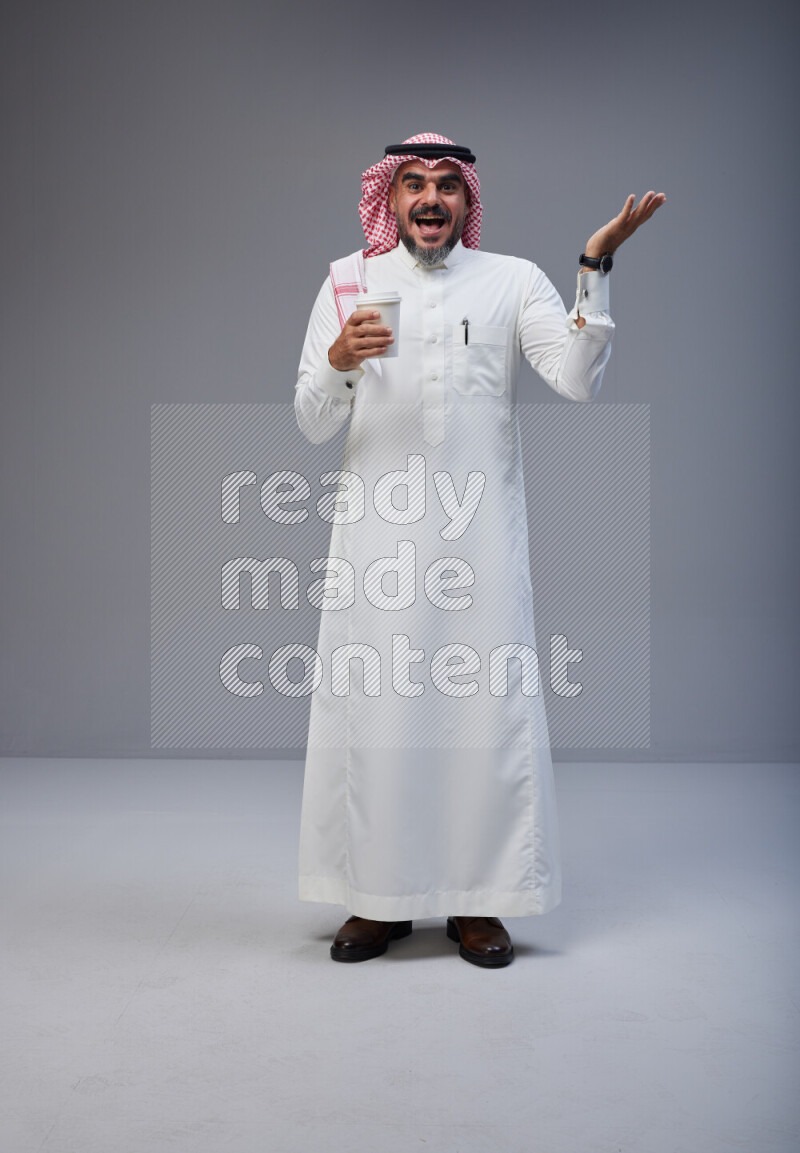Saudi man Wearing Thob and red Shomag standing holding paper cup on Gray background