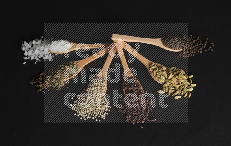 wooden spoons filled with white peppers, cloves, cardamom, salt, black peppers and basil on black flooring