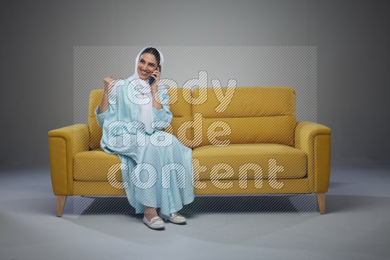 A Saudi woman wearing a light blue Abaya and a white head scarf sitting on a yellow sofa and talking in the phone while making different poses eye level on a grey background